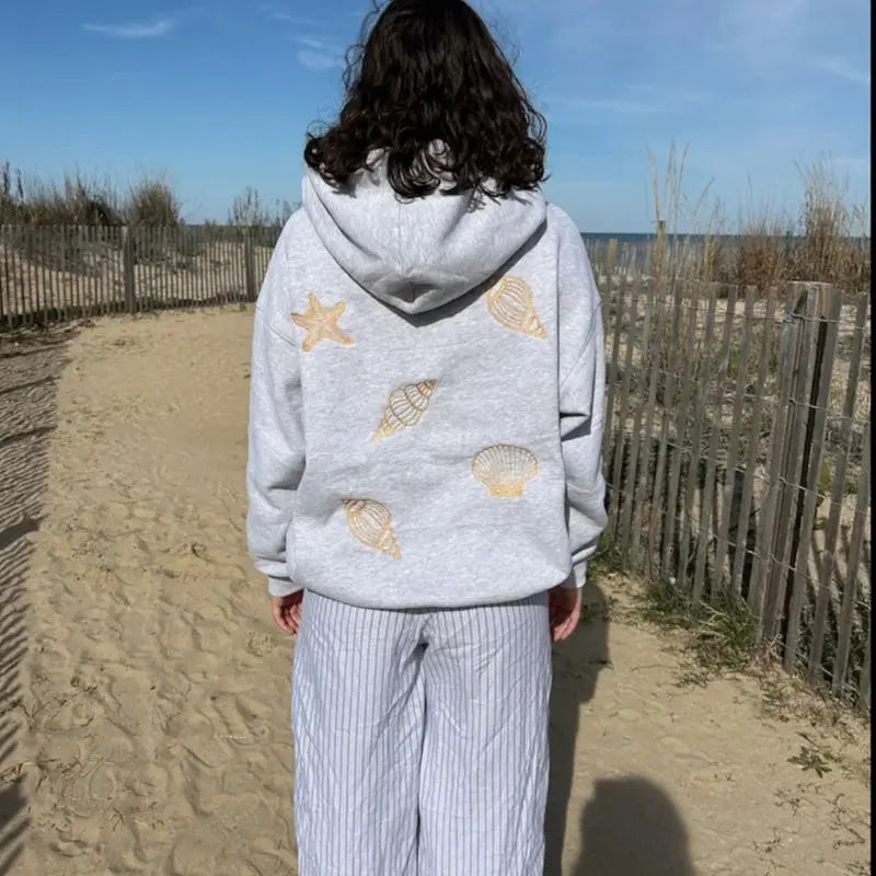 Person wearing a gray hoodie with shell patterns on a sandy path by the beach.