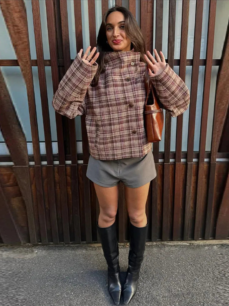 Woman wearing a plaid jacket, gray shorts, and black knee-high boots standing against a wooden fence.
