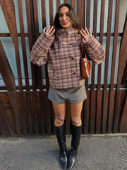 Woman wearing a plaid jacket, gray shorts, and black knee-high boots standing against a wooden fence.