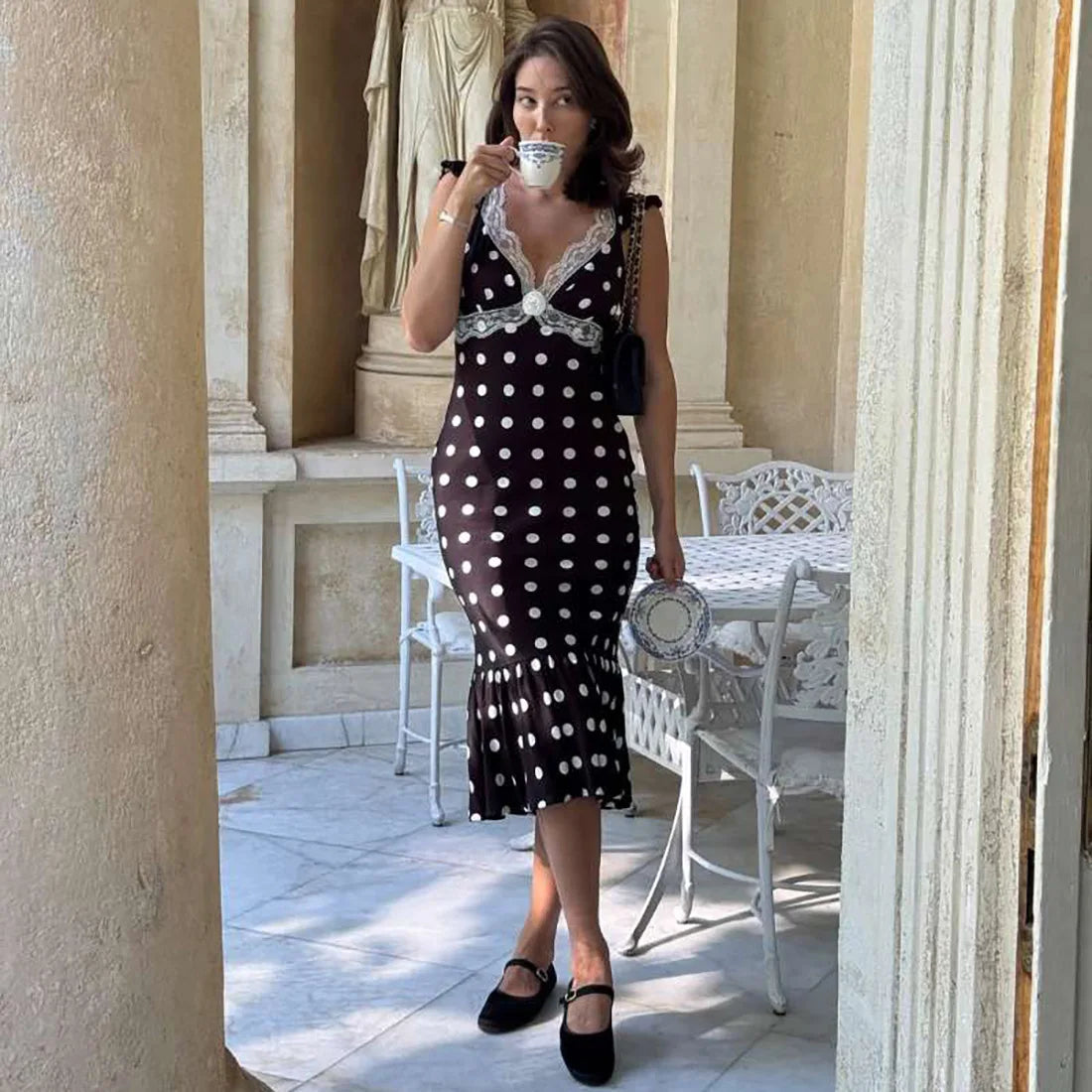 Woman in a black and white polka dot dress drinking from a teacup in an elegant outdoor setting.