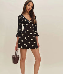 Woman wearing a black dress with white polka dots holding a brown handbag on a beige background