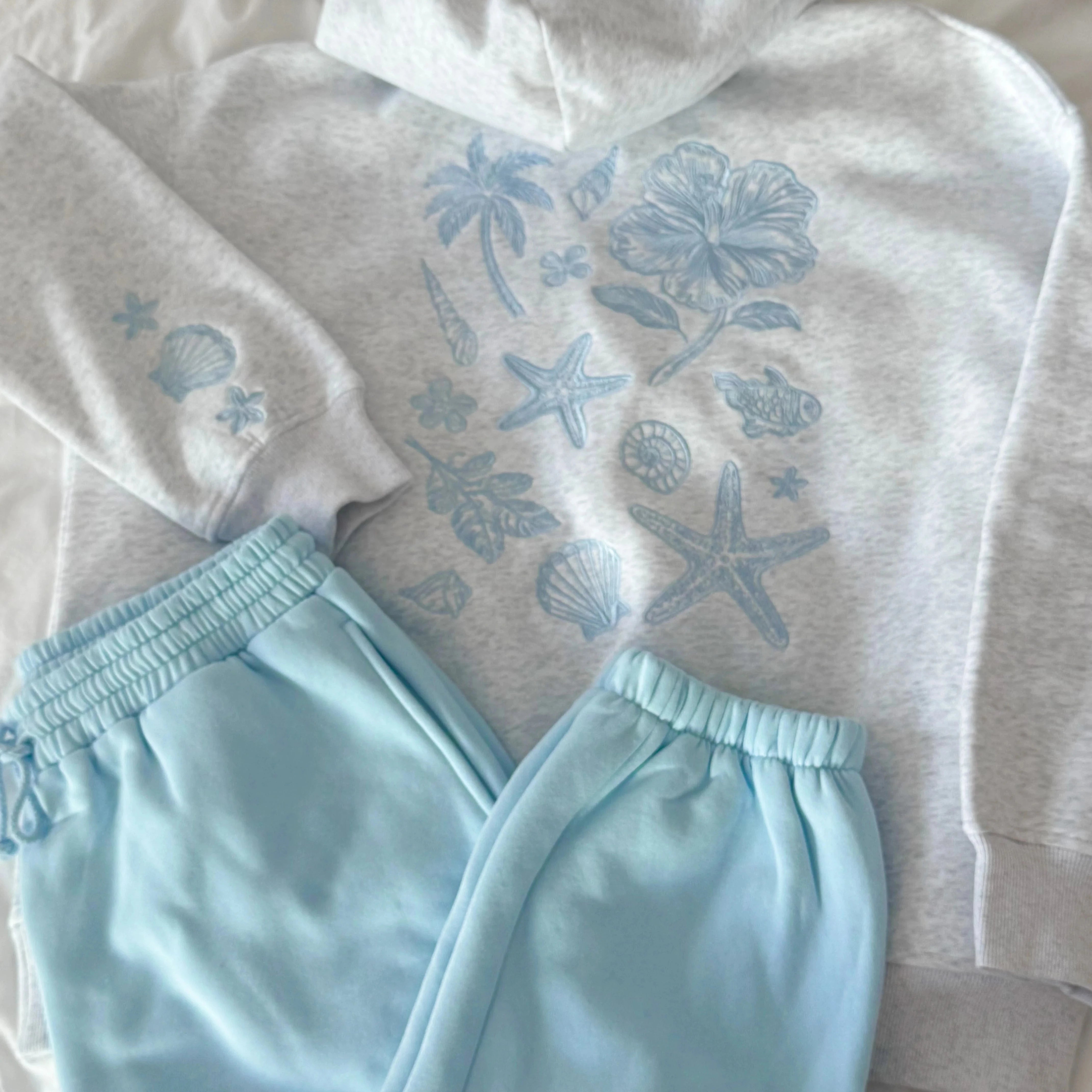 Light gray hoodie with blue ocean-themed designs and matching light blue pants on a neutral background