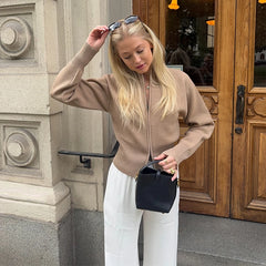 Woman in beige sweater and white pants holding a black handbag, standing in front of a wooden door.