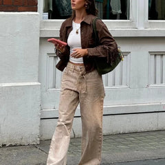 Woman in brown jacket and beige pants holding a pink phone, standing on a street.