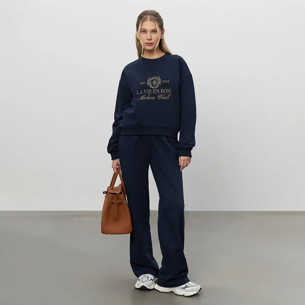 Woman wearing a navy blue sweatshirt and pants with a brown handbag on a plain background