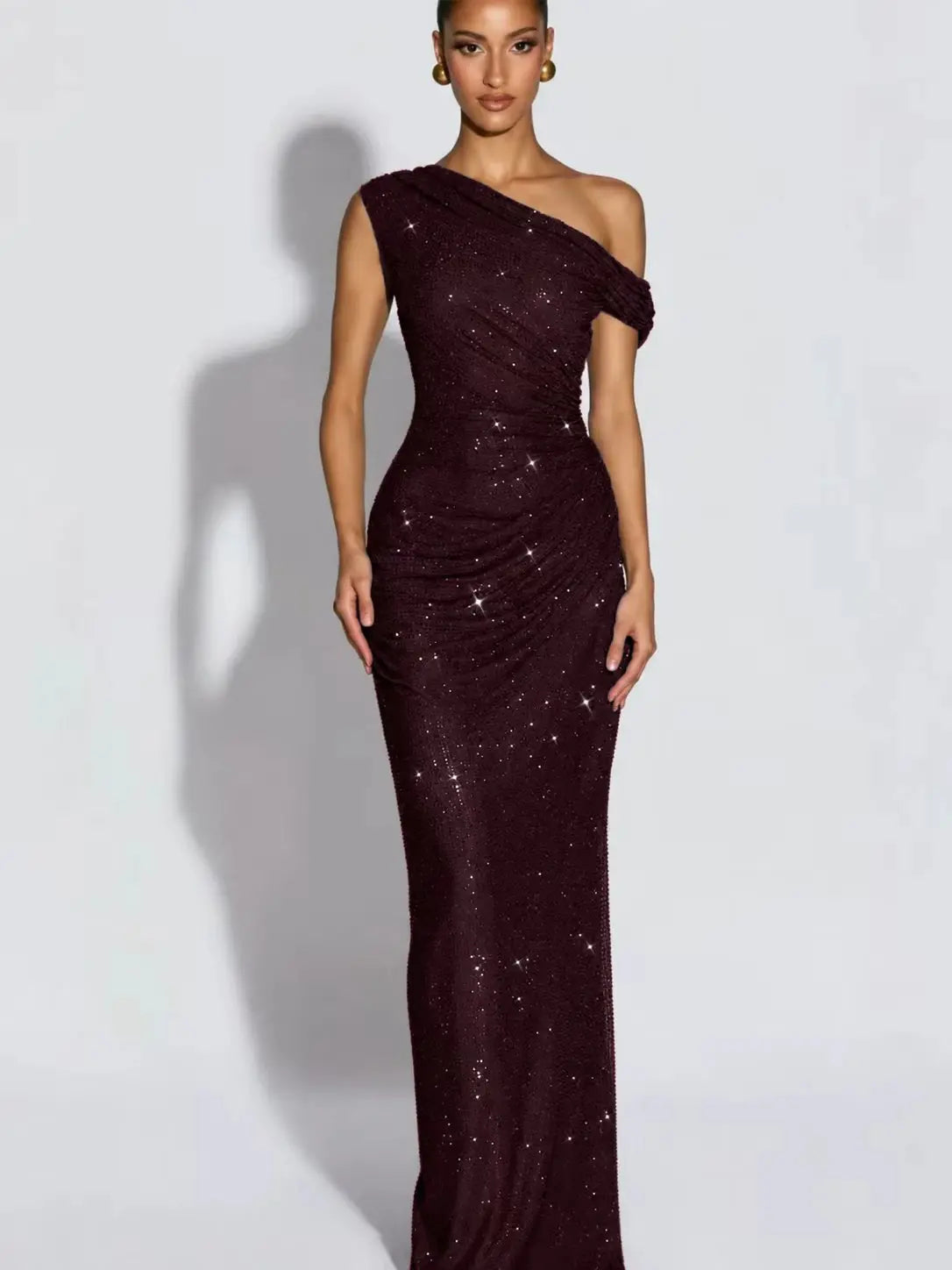 A woman modeling a floor-length, one-shoulder evening gown covered in sparkling sequins. The dress is a wine color and is photographed against a plain light gray studio background with a soft shadow cast to the side.