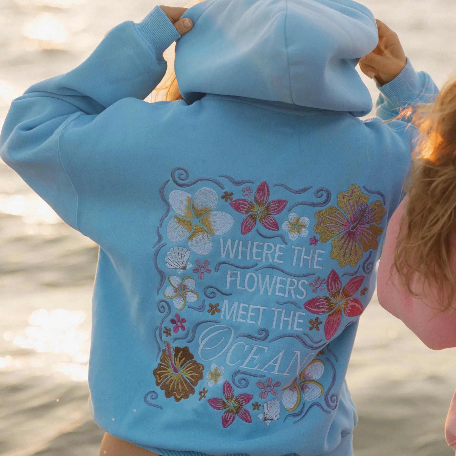 Person wearing a blue hoodie with floral designs and text on a beach
