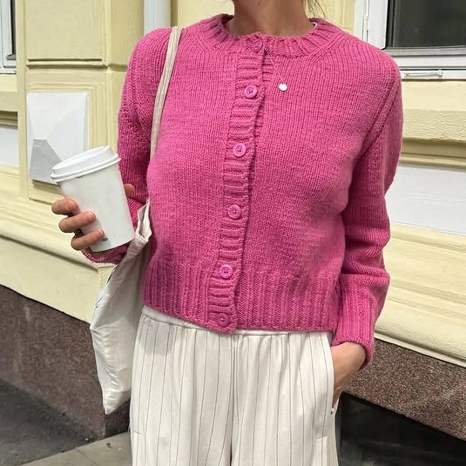 Person wearing a pink knitted cardigan holding a white cup outdoors.