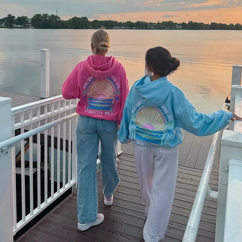 Two people standing on a dock by a lake, wearing colorful hoodies with artistic designs.