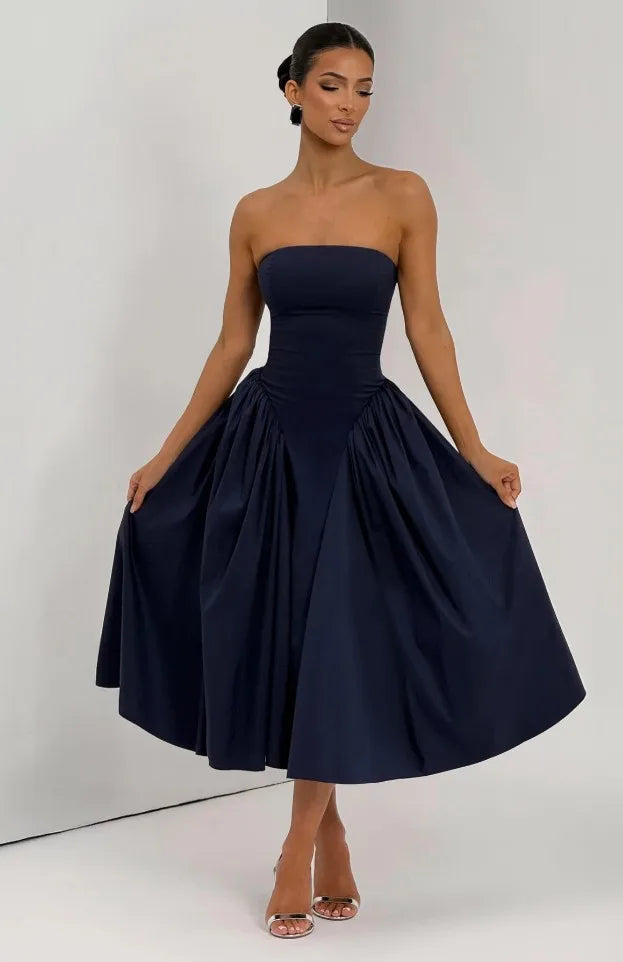 A woman modeling a strapless navy blue midi dress with a structured corset-style bodice and a full, gathered skirt, standing against a plain light gray studio background.