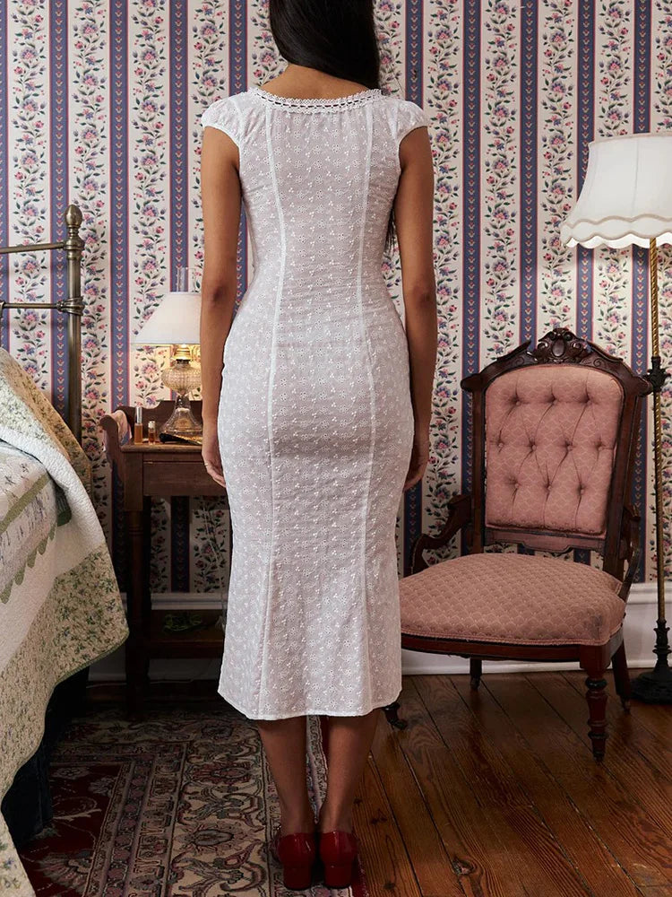 Woman in a white dress standing in a room with floral wallpaper and wooden furniture.