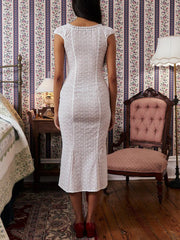 Woman in a white dress standing in a room with floral wallpaper and wooden furniture.