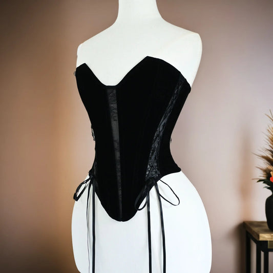 A black velvet corset with sheer mesh panels and side laces, displayed on a white mannequin torso against a warm beige wall.