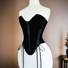 A black velvet corset with sheer mesh panels and side laces, displayed on a white mannequin torso against a warm beige wall.