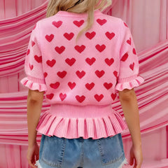 Person wearing a pink sweater with red heart patterns in front of a pink background