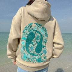Person wearing a beige hoodie with teal ocean-themed design on a beach