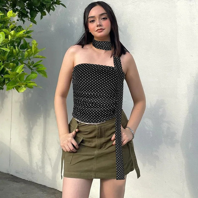 Woman wearing a black polka dot top and green skirt with a white wall and plant in the background