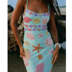 Woman wearing a blue dress with colorful shell and starfish patterns on a beach.