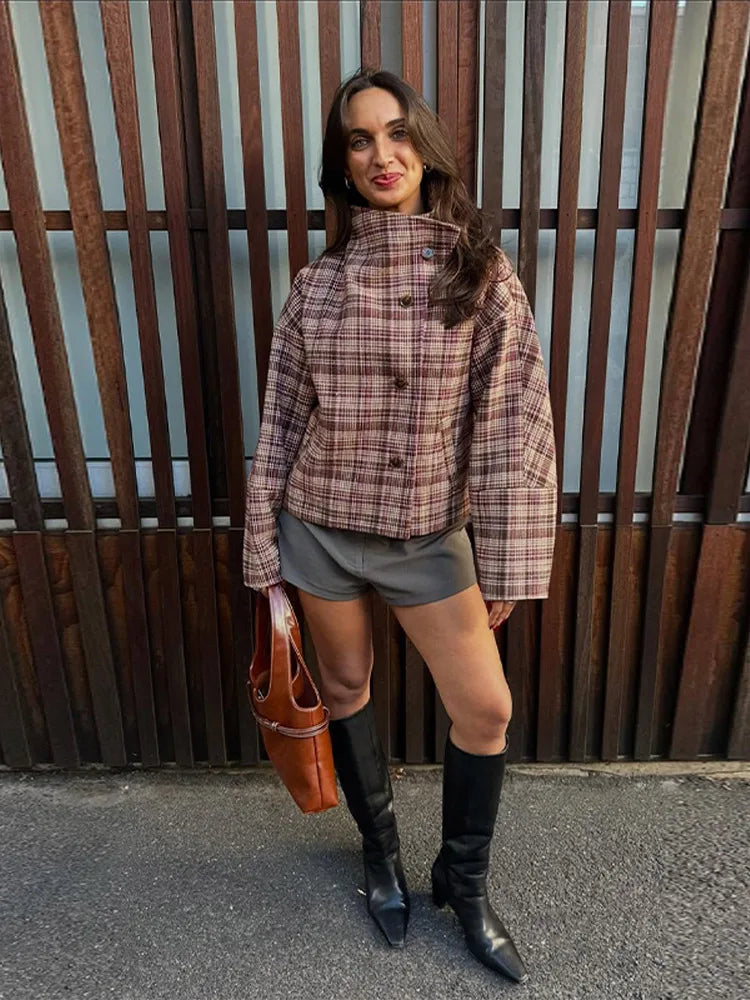 Woman wearing a plaid shirt, shorts, and knee-high boots standing against a wooden fence.