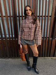 Woman wearing a plaid shirt, shorts, and knee-high boots standing against a wooden fence.