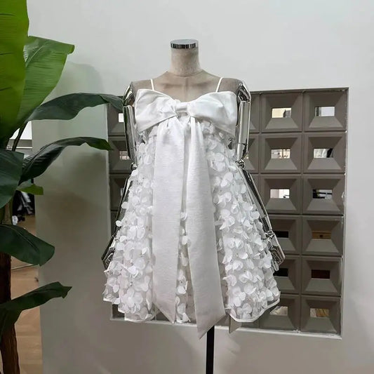 White mini dress with spaghetti straps, oversized satin bow detail on the bodice, and a sheer skirt adorned with 3D floral appliqués and sequins, displayed on a mannequin.