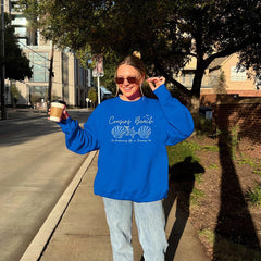 Person wearing a blue sweatshirt with text and graphics, holding a coffee cup on a sidewalk.