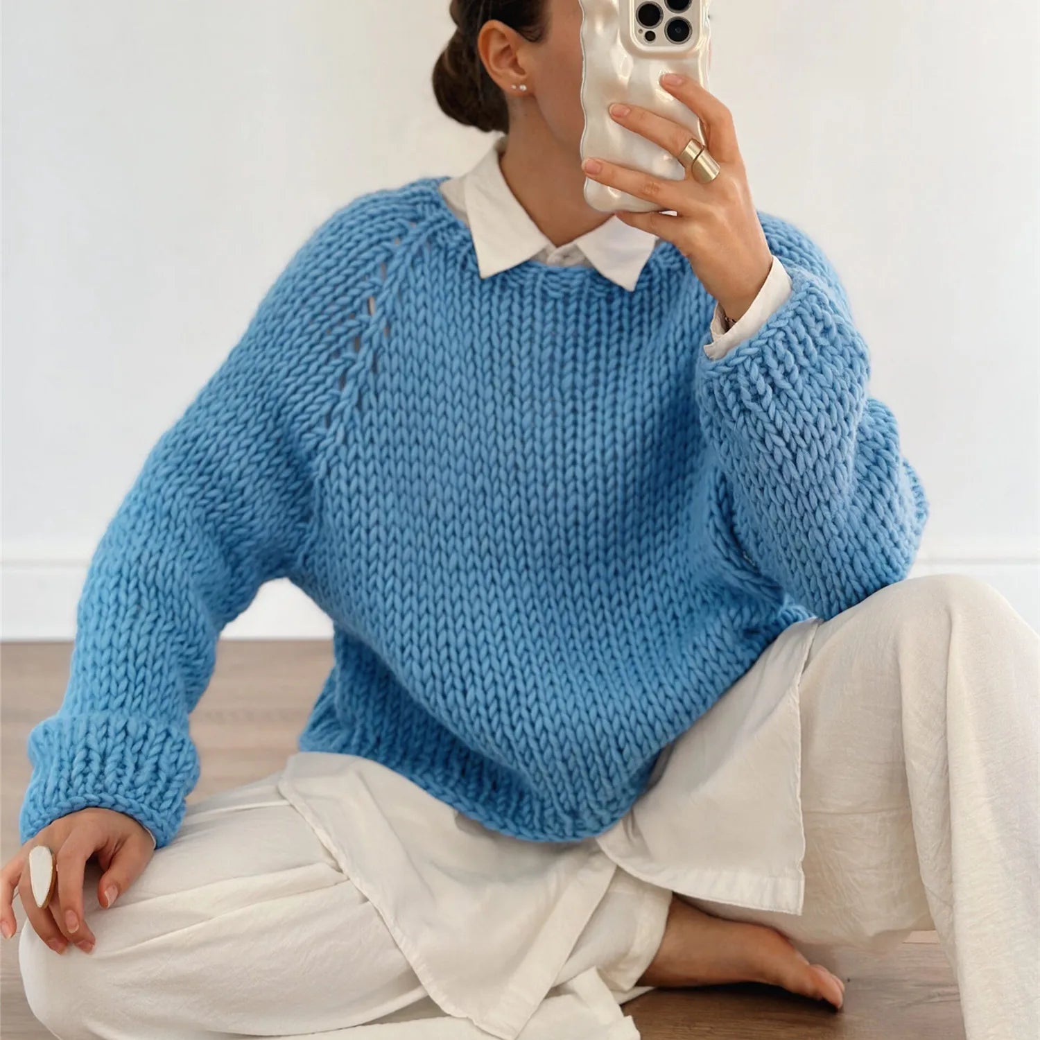 Person wearing a blue knitted sweater with white shirt and pants, sitting on a wooden floor.