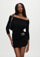 A woman modeling a black off-the-shoulder long-sleeve mini dress with a silver rectangular belt buckle at the waist, paired with a black shoulder bag, against a plain light gray studio background.
