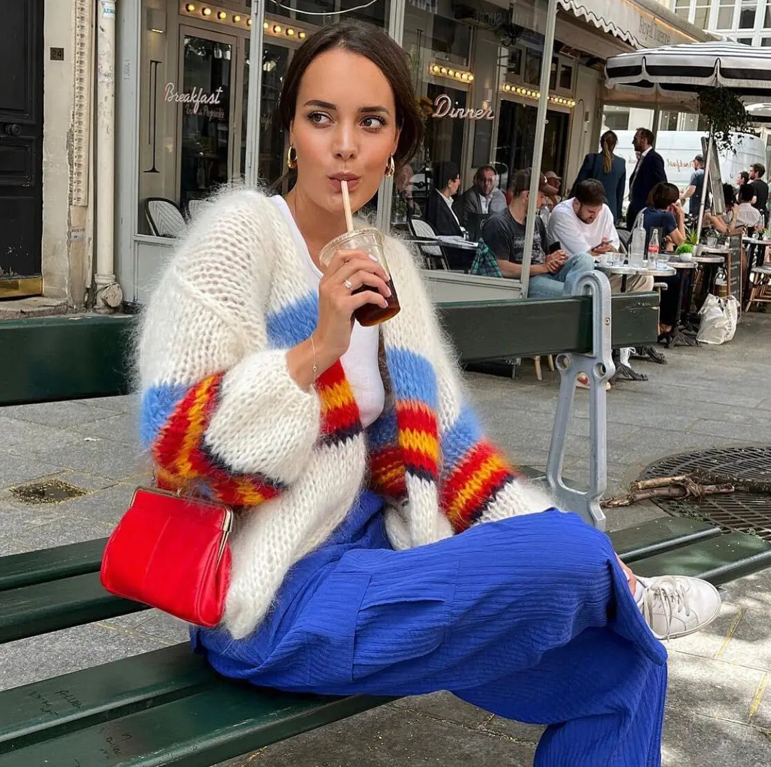 Woman sitting on a bench wearing a colorful sweater and blue pants, holding a drink.
