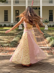 A woman with long brown hair is seen from behind, twirling in a pastel pink and yellow floral two-piece outfit with sheer panels, set against a backdrop of a white colonial-style house and brick pathway.