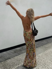 Woman in a floral dress with a black bag, arms outstretched, against a plain wall.
