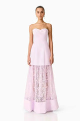 A woman modeling a strapless light pink gown with a structured sweetheart bodice and a full skirt featuring sheer floral lace overlay, photographed against a plain white studio background.
