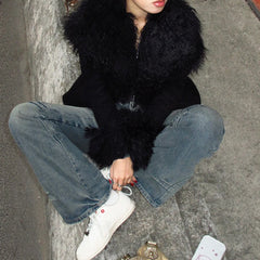 Person wearing a black fur-lined coat, blue jeans, and white sneakers sitting on a concrete surface.