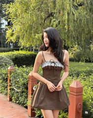 A woman wearing a brown mini dress with a silver patterned bustle strapless bodice stands outdoors in a park-like setting with green hedges and trees.