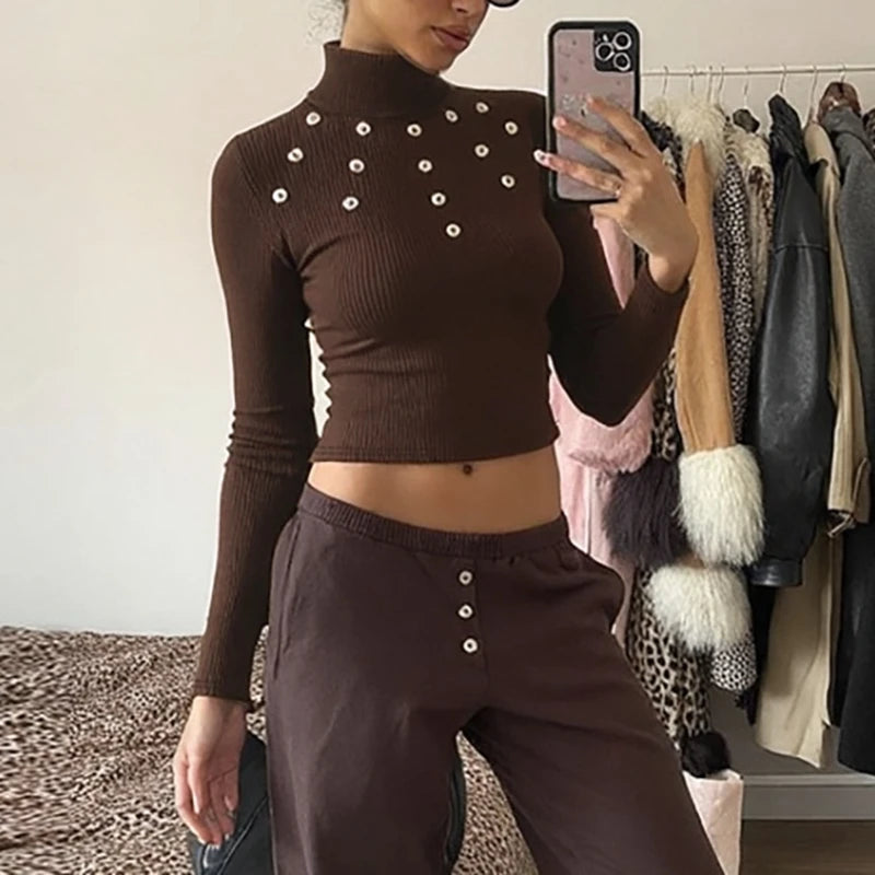 Person wearing a brown ribbed turtleneck and pants with button details, standing in front of a clothing rack.