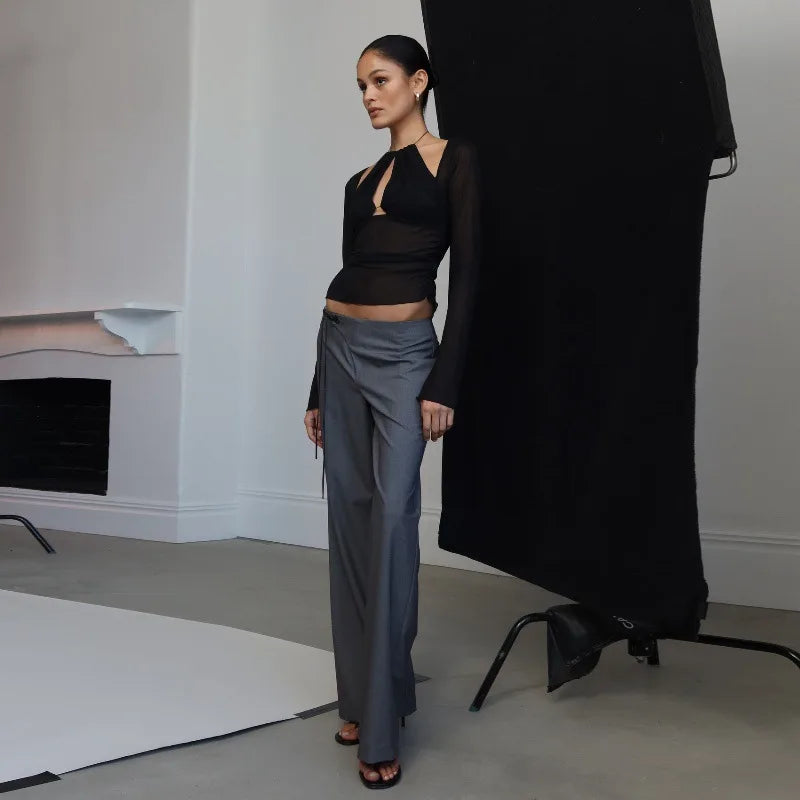 Woman in black top and gray pants standing in a minimalistic room with a black coat draped over a chair.