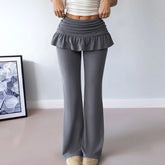 Person wearing gray flared pants with a ruffled top against a white wall.