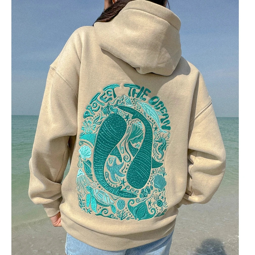 Beige hoodie with teal ocean-themed design on a beach