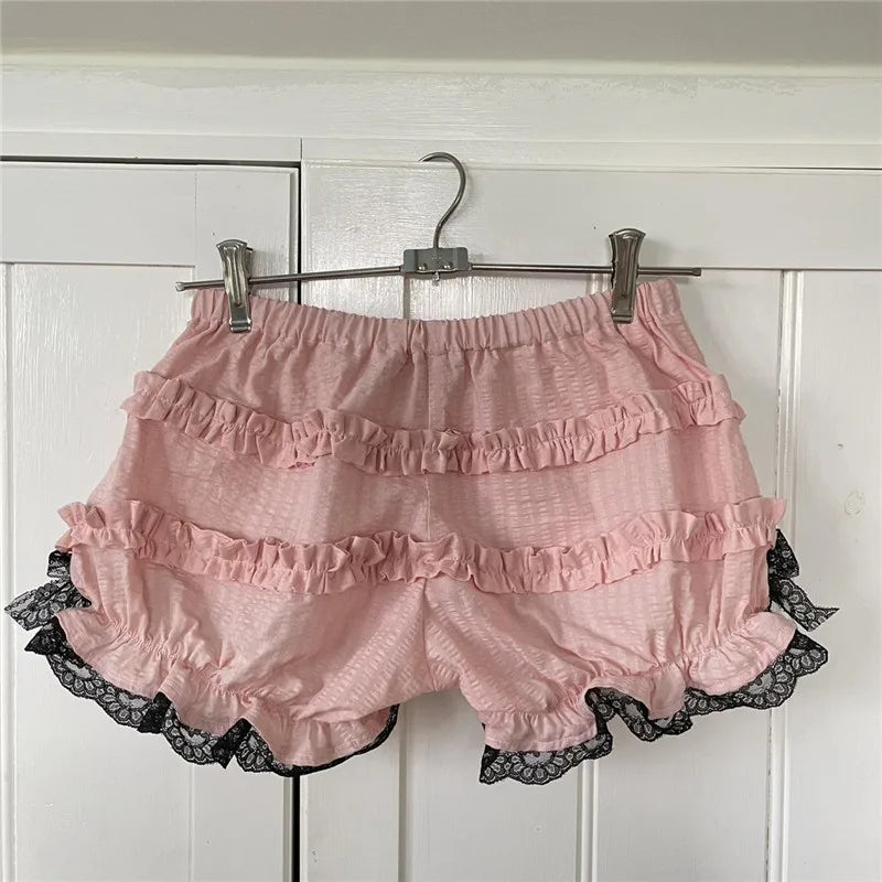 Pink ruffled shorts with black lace trim hanging on a rack against a white wooden panel background.