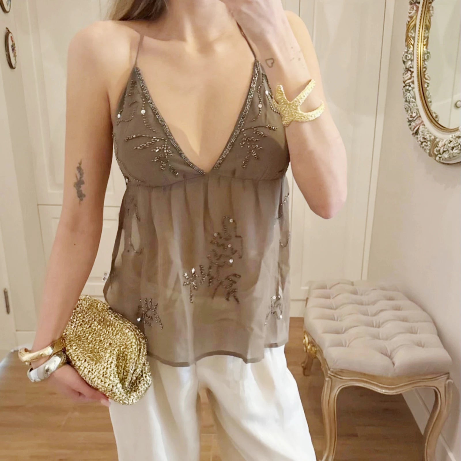 Woman wearing a sheer top with embroidery and holding a gold clutch in a room.