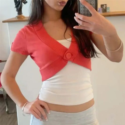 Model wearing a coral cropped knit bolero over a white tank top, hands on hips.