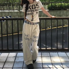 Person wearing a white t-shirt and beige pants with text, standing on a tiled floor.