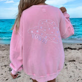 Person wearing a pink sweatshirt with 'HOWDY' and a hat graphic on a beach.