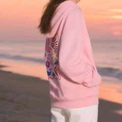 Person wearing a pink hoodie with colorful designs on a beach at sunset