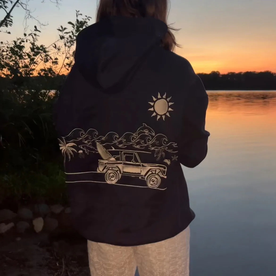 Person wearing a hoodie with a graphic design of a beach scene at sunset by a lake.