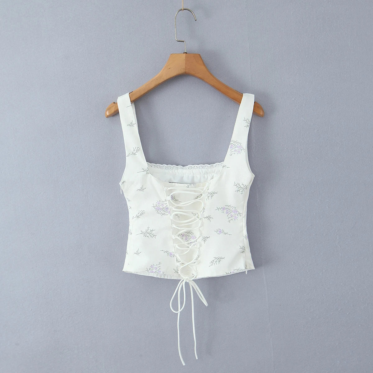 White floral lace-up crop top on a wooden hanger against a gray background