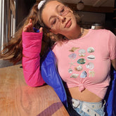 Person wearing a pink shirt with shell designs, blue jacket, and denim shorts outdoors.