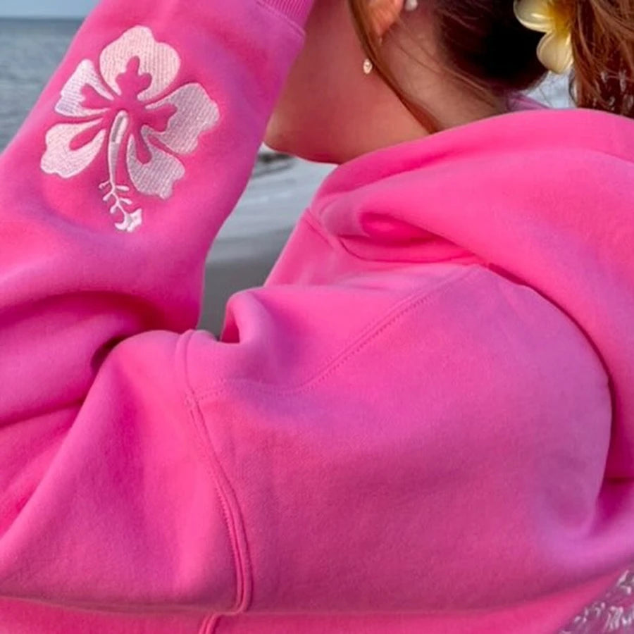 Person wearing a bright pink hoodie with a white floral design on the sleeve, standing by a body of water.
