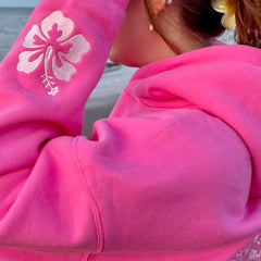 Person wearing a bright pink hoodie with a white floral design on the sleeve, standing by a body of water.