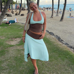 Woman wearing a light blue and brown two-piece set with a halter crop top and asymmetrical midi skirt, posing on a grassy beach area.